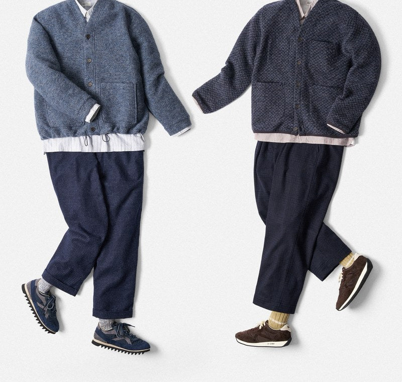 Twee platgelegde outfits van Universal Works: wollen vesten in donker- en lichtblauw, wit overhemd, blauwe broek en sneakers.